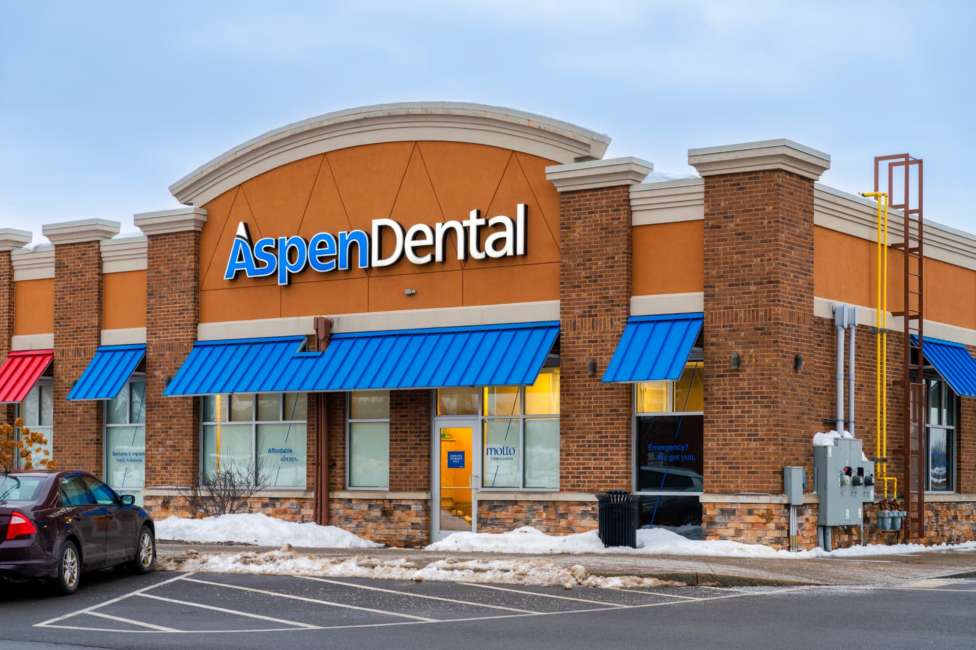 Exterior of an Aspen Dental office with blue and orange signage and dentures and implants window messaging, shown in an Aspen Dental implants review