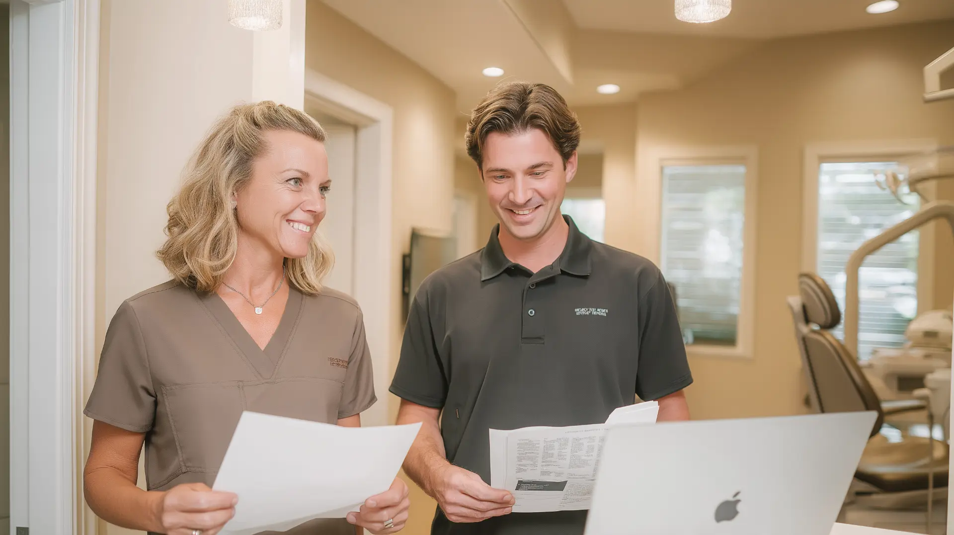Dentist and office manager standing in a bright clinic hallway, each holding a printed marketing report with columns of numbers, reviewing them together at a laptop, the one page report every dental marketing agency should send