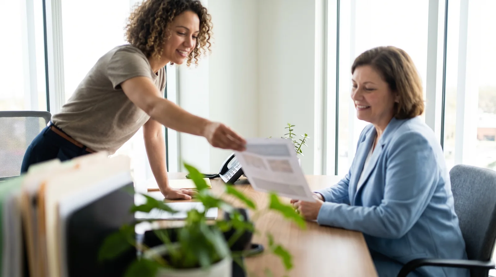 Coordinator handing a patient a printed one-page comparison, the page out of focus, both seated at a desk