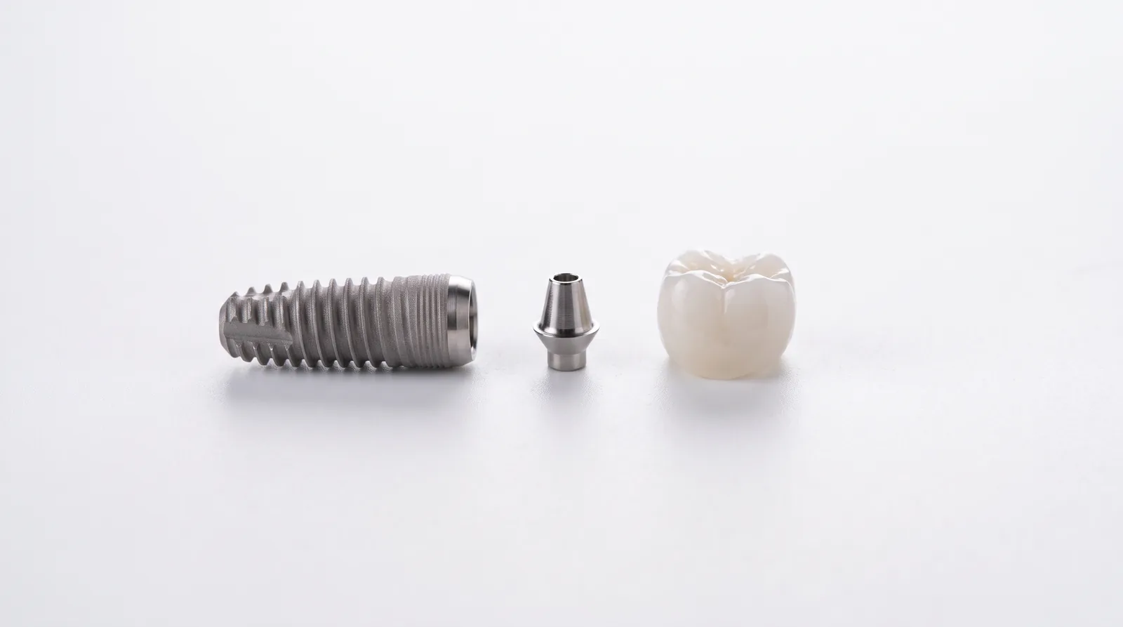 Single dental implant fixture, abutment, and ceramic crown laid out in a row on a white surface with soft shadows