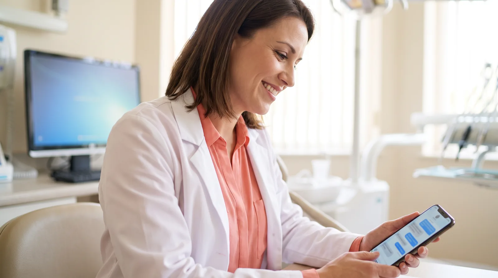 A treatment coordinator replying to a patient's Messenger question about All-on-4 pricing on a phone, with a Facebook ad for the practice visible on a monitor behind