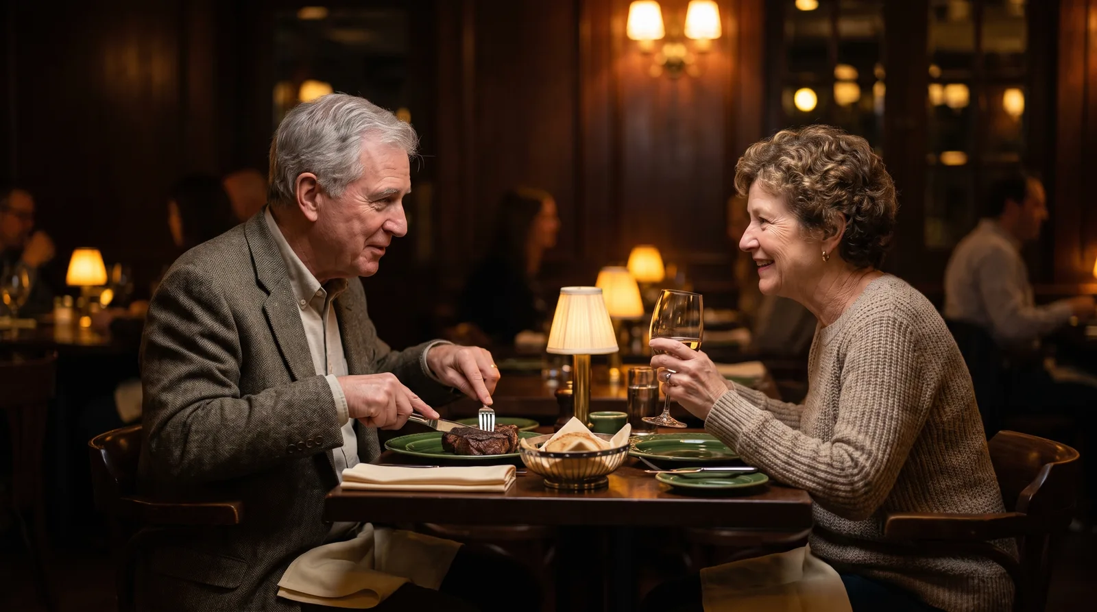 Older man and woman eating a normal dinner at a restaurant table, one cutting into steak, both faces visible, warm evening light