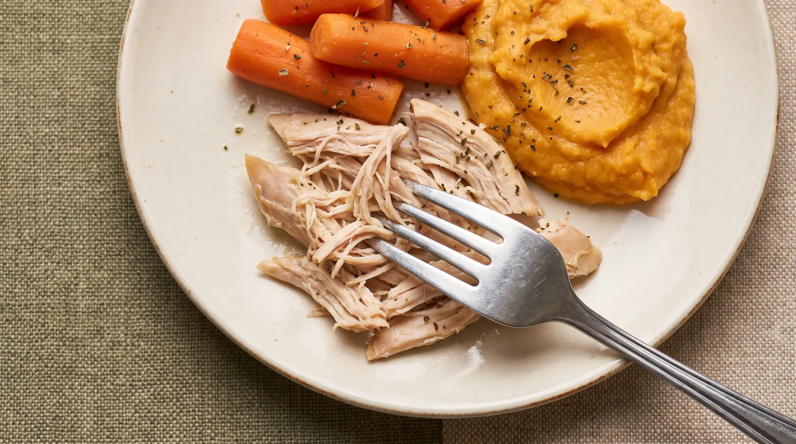 Fork breaking apart slow-cooked shredded chicken with steamed carrots on a plate, overhead view, no hands in frame